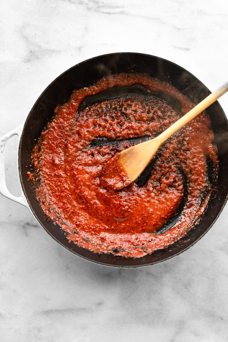 Thick red sauce cooking in a skillet, being stirred with a wooden spoon.