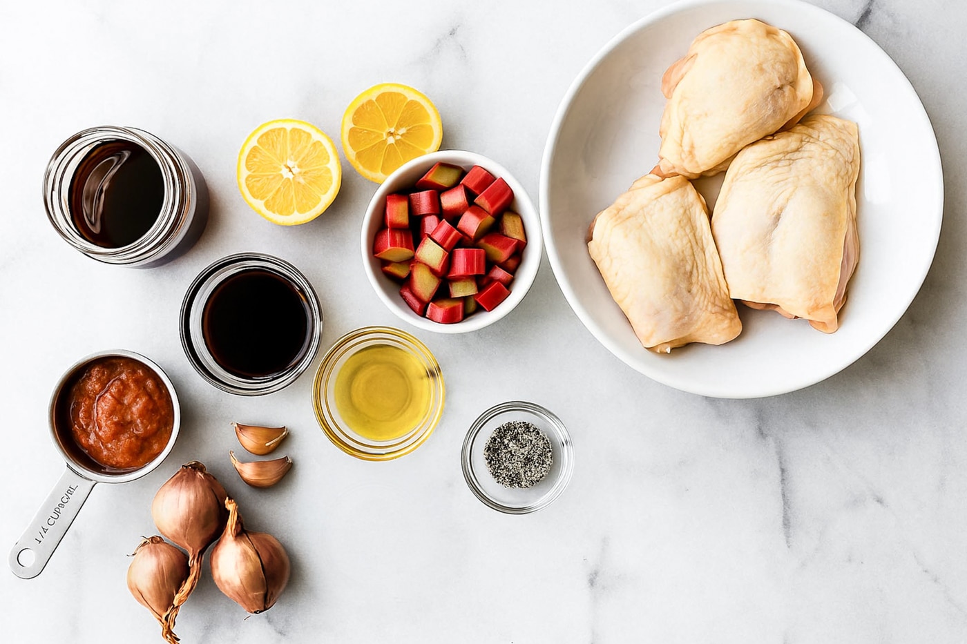 Ingredients laid out on a counter including raw chicken thighs, chopped rhubarb, lemon, shallots, oil, and seasonings.