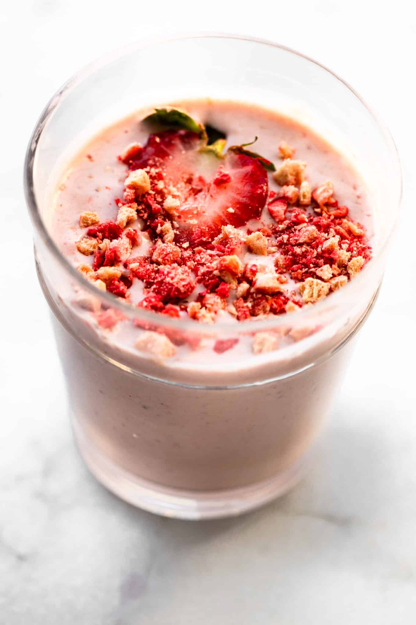 Close-up of a creamy pink strawberry smoothie in a clear glass, topped with fresh strawberry slices and crunchy strawberry crumble on a marble countertop.