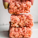Hand lifting a strawberry rice cereal treat from a stack of three pink cereal bars.