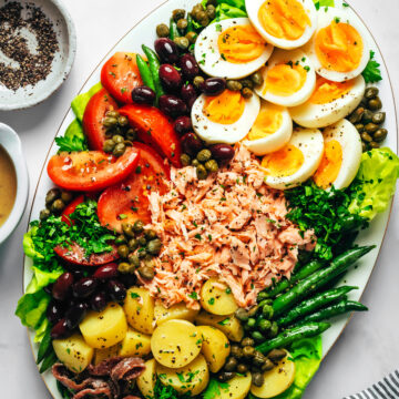 A salmon nicoise salad with salad greens, green beans, new potatoes, tomatoes, hard boiled eggs, olives, capers, salmon and herbs on an oval platter.
