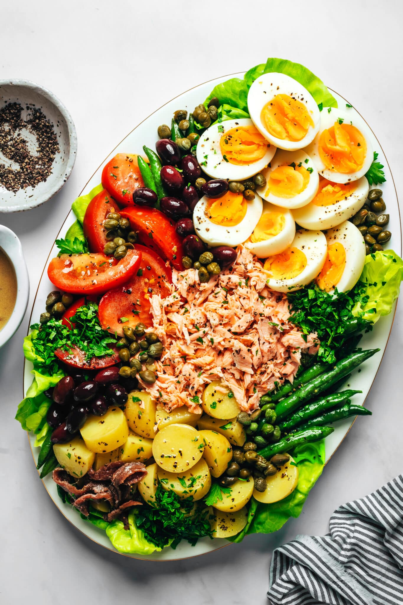 A salmon nicoise salad with salad greens, green beans, new potatoes, tomatoes, hard boiled eggs, olives, capers, salmon and herbs on an oval platter.