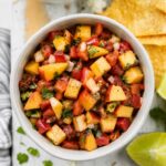 small white bowl full of peach salsa on white wood board with chips and a lime wedge around it
