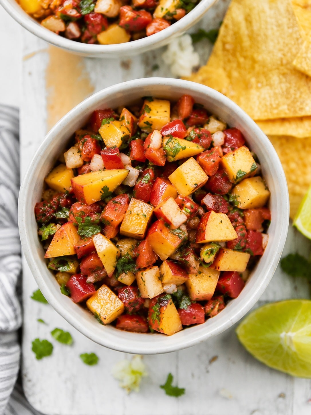 small white bowl full of peach salsa on white wood board with chips and a lime wedge around it