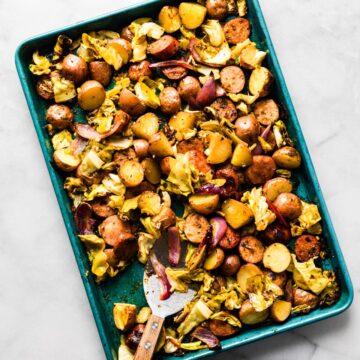 Large sheet pan of roasted cabbage, sausage, and crispy potatoes with spatula resting in it.